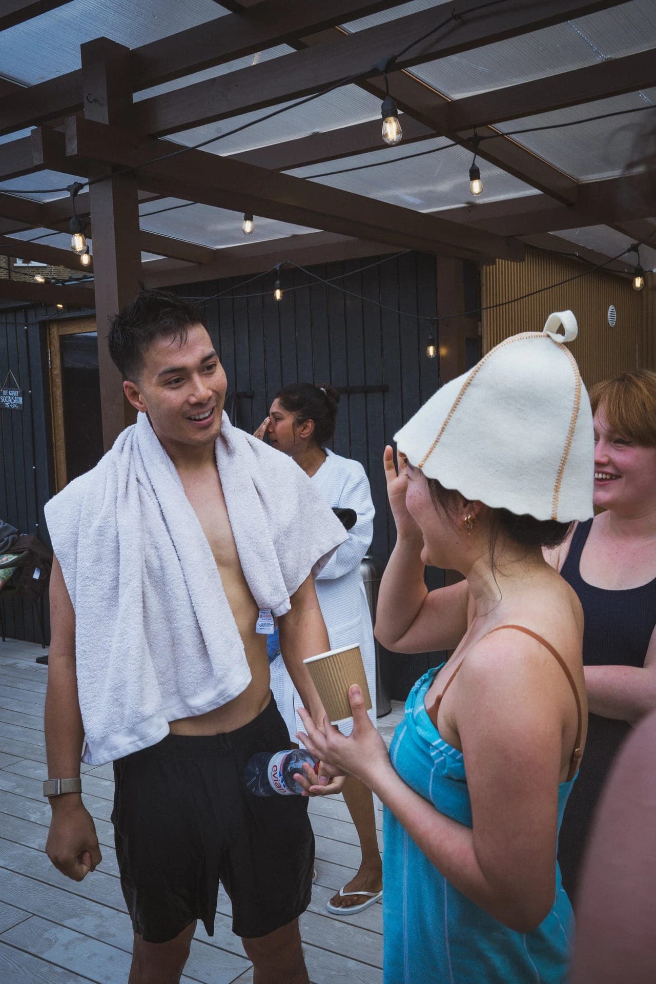 Friends laughing on the deck between sauna rounds, one in a felt sauna hat